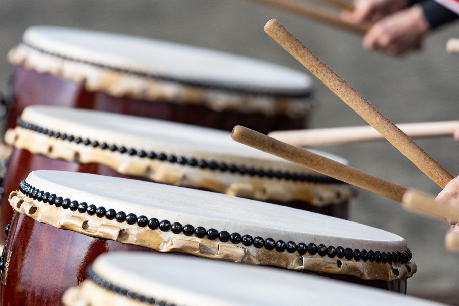 People holding and beating wood drumsticks on row of drums